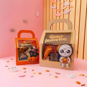 Puede incluir: Dos bolsas de regalo de cartón con asas y ventanas transparentes. La bolsa naranja pequeña muestra un gato negro sobre una calabaza. La bolsa beige grande muestra una momia con una calabaza. Ambas bolsas tienen el texto Happy Halloween.