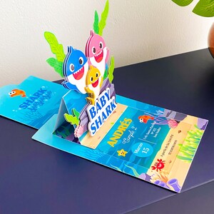 K&ouml;nnte beinhalten: Eine blaue, gelbe und rosafarbene Baby Shark Pop-up-Karte als Einladung zu einem zweiten Geburtstag eines Kindes. Die Karte zeigt den Text "Baby Shark" und "Andr&eacute;s Cumple 2" mit dem Datum 15. M&auml;rz.