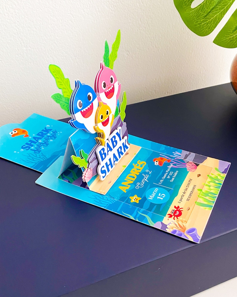 K&ouml;nnte beinhalten: Eine blaue, gelbe und rosafarbene Baby Shark Pop-up-Karte als Einladung zu einem zweiten Geburtstag eines Kindes. Die Karte zeigt den Text "Baby Shark" und "Andr&eacute;s Cumple 2" mit dem Datum 15. M&auml;rz.