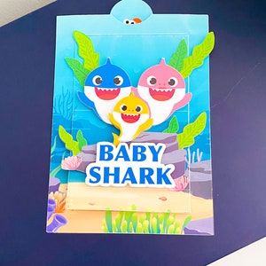 K&ouml;nnte beinhalten: Eine blaue, rosa und gelbe Baby Shark Familienkarte mit den Worten "Baby Shark" in blauen Buchstaben.