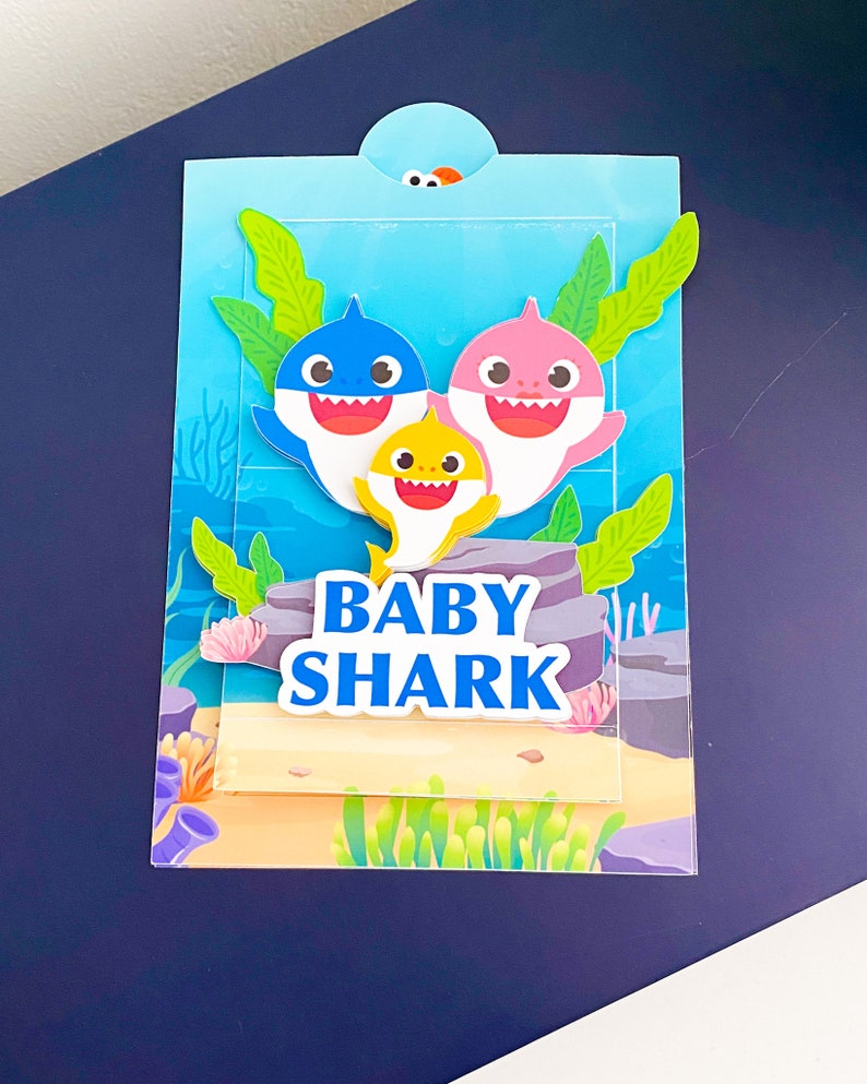 K&ouml;nnte beinhalten: Eine blaue, rosa und gelbe Baby Shark Familienkarte mit den Worten "Baby Shark" in blauen Buchstaben.