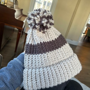 May include: A knitted beanie with a pom pom on top. The beanie is white with brown stripes.