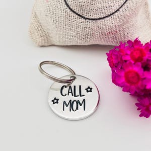 Puede incluir: Un llavero redondo de metal plateado con las palabras "CALL MOM" en escritura negra, acentuado con formas de estrella. El llavero está unido a un anillo plateado. Un ramo de flores rosa brillante y una bolsa beige están en el fondo.