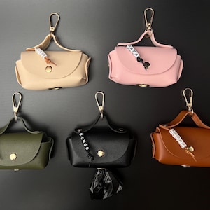 Op de afbeelding: Vijf kleine, rechthoekige poepzakjeshouders van kunstleer in beige, roze, olijfgroen, zwart en bruin. Elk exemplaar heeft een goudkleurige drukknoopsluiting, een handvat met een goudkleurige karabijnhaak en een gepersonaliseerde kralenbedel.