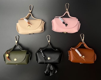 May include: Five small, rectangular, faux leather waste bag dispensers in beige, light pink, olive green, black, and brown. Each features a gold-tone snap closure, a top handle with a gold-tone lobster clasp, and a personalized bead charm.