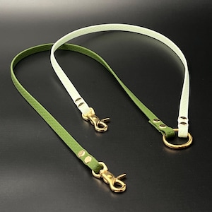 Op de afbeelding: Twee leren riemen, één groen en één wit, met goudkleurige metalen sluitingen en hardware. De riemen zijn dun en flexibel, waarschijnlijk voor een tas of accessoire. De achtergrond is effen zwart.
