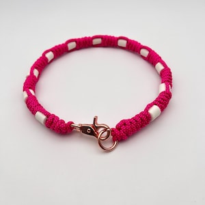Pode incluir: Uma pulseira circular feita de cordão trançado em rosa choque, com contas cilíndricas brancas espaçadas uniformemente. As extremidades são presas com um fecho lagosta de metal em ouro rosa e um anel circular correspondente.