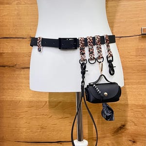 Pode incluir: Um cinto preto com fivela quadrada e detalhes de estampa de leopardo. O cinto tem vários acessórios, incluindo uma pequena bolsa preta com um botão dourado e uma longa alça preta. O cinto é exibido em um manequim branco.