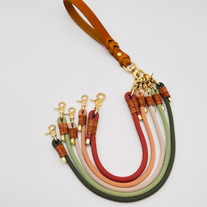 Puede incluir: Un llavero multicolor con una correa de muñeca de cuero marrón y herrajes dorados. El llavero presenta seis hebras de cuerda en verde, naranja, rojo, beige y negro. Cada hebra tiene una sección decorativa envuelta.