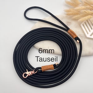 Pode incluir: Uma trela de corda preta com um mosquetão giratório de metal em ouro rosa e enrolamentos de cordão castanho-claro nas extremidades da pega e do clipe. A trela está enrolada em círculo e inclui o texto 6mm Tauseil impresso no centro.