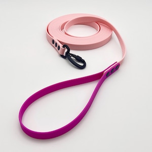 Pode incluir: Uma trela comprida para cão com um design de dois tons, apresentando uma secção enrolada em rosa claro e uma pega em magenta vibrante. A trela é feita de um material liso e flexível, incluindo um mosquetão giratório de metal preto e rebites.