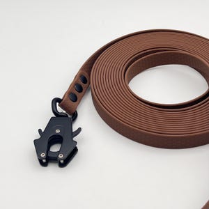 Pode incluir: Uma trela para cão castanha com um padrão hexagonal texturado e um fecho tático de metal preto mate. A trela está enrolada numa forma circular e apresenta três rebites pretos que fixam a fita ao mecanismo de gancho giratório resistente.