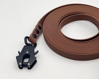 Op de afbeelding: Een bruine hondenriem met een textuur van zeshoekige patronen en een matzwarte tactische metalen sluiting. De riem is in een cirkelvorm opgerold en is voorzien van drie zwarte klinknagels die de band aan het robuuste draaibare haakmechanisme bevestigen.