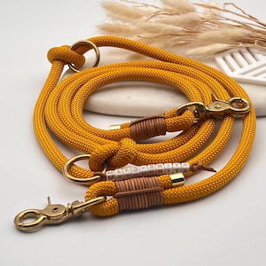 Op de afbeelding: Een mosterdgele hondenriem van touw met messing hardware, inclusief twee draaibare musketonhaken en twee ringen. De riem is afgewerkt met bruine leren wikkelingen en een gepersonaliseerd gedeelte met witte vierkante kralen die MARGHERITA spellen.