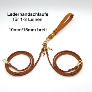 Pode incluir: Uma alça de guia de couro castanho com um ponto de conexão trançado, apresentando clipes giratórios de metal dourado e dois segmentos de guia enrolados. O texto diz: Lederhandschlaufe für 1-3 Leinen, 10mm/16mm breit.