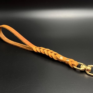 Könnte beinhalten: Eine geflochtene Lederhundeleine mit einem goldenen Metallclip. Die Leine ist hellbraun.
