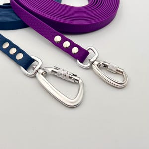 Op de afbeelding: Twee lange, platte lijnen in donkerblauw en paars, elk voorzien van een gestructureerd honingraatpatroon. Beide lijnen zijn vastgezet met zilverkleurige metalen klinknagels en hebben een stevige, vergrendelbare zilveren karabijnhaak.
