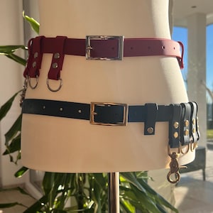 Könnte beinhalten: Zwei Gürtel an einem Schaufensterpuppen-Torso. Der obere Gürtel ist dunkelrot mit einer silberfarbenen Schnalle und zwei silberfarbenen Ringen. Der untere Gürtel ist schwarz mit einer goldfarbenen Schnalle und mehreren schwarzen Schlaufen mit Gold.