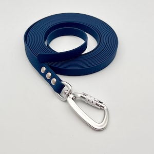 Pode incluir: Uma trela para cão azul-escura enrolada em forma circular, com uma superfície texturizada e um mosquetão de metal prateado fixado com três rebites prateados. A trela é feita de um material flexível com um acabamento mate e liso.
