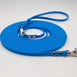 Pode incluir: Uma trela longa de cor azul brilhante feita de um material liso e flexível. A trela possui uma alça numa extremidade, ganchos giratórios de metal prateado em ambas as extremidades e é fixada com rebites de metal prateado nas junções.