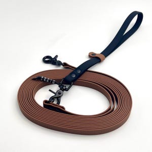 Pode incluir: Uma trela para cão castanha com pega preta e ferragens em metal preto. A trela inclui um segundo gancho preto e um pequeno pendente preto com contas brancas que formam o nome GIBBS. A fita tem uma superfície texturada e está enrolada.