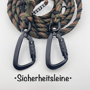 Op de afbeelding: Een gevlochten paracord-lijn met een camouflagepatroon in olijfgroen, bruin en zwart. De lijn is voorzien van twee matzwarte metalen karabijnhaken met schroefsluiting en een kleine kraal met de tekst HUNTER.