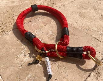 Op de afbeelding: Een rode hondenhalsband van touw met zwarte koordwikkelingen, goudkleurige metalen ringen en een bedeltje met de naam ROCKY op witte kubuskralen. De halsband is gemaakt van gevlochten rood nylontouw met zwarte accenten.