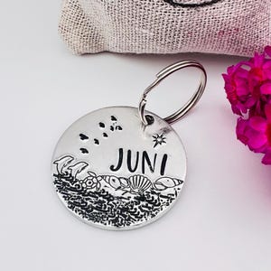 Può includere: Un portachiavi rotondo color argento con inciso il nome "JUNI". Il tag presenta una scena di spiaggia con delfini, una tartaruga, conchiglie e una stella. Un portachiavi argentato è attaccato. Un fiore rosa e un sacco di iuta sono sullo sfondo.