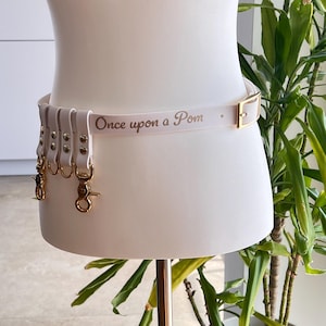 Può includere: Una cintura bianca con il testo color oro "Once upon a Pom" stampato sulla fascia. La cintura include una fibbia color oro e diversi passanti verticali bianchi, ciascuno fissato a un moschettone girevole in metallo dorato per appendere oggetti.