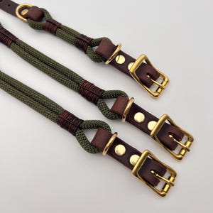Op de afbeelding: Een set olijfgroene touwriemen met donkerbruine lederen accenten en goudkleurige metalen hardware. De riemen zijn gemaakt van gevlochten koord, vastgezet met lederen wikkelingen, en bevatten verstelbare gespen en D-ringen voor bevestiging.
