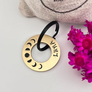 Puede incluir: Un llavero de metal dorado con un llavero negro. El llavero presenta un diseño de fases lunares y la palabra "Luna" en una fuente manuscrita. Una pequeña bolsa está en el fondo, y flores de color rosa brillante están a la derecha.