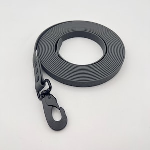 Pode incluir: Uma trela impermeável cinzento-escura enrolada em forma circular, com um mosquetão giratório de metal preto mate. A trela é feita de um material liso e flexível com uma superfície texturizada, fixada com três rebites cinzento-escuros a condizer.