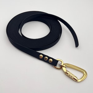 Pode incluir: Uma trela longa, preta e plana enrolada em forma circular. A trela é feita de um material liso e mate, com três rebites metálicos dourados que prendem a fita a um grande mosquetão metálico dourado com um mecanismo de bloqueio.