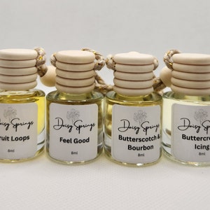 May include: Four small glass bottles with wooden stoppers and string hangers. Each bottle contains a yellow liquid and has a white label with black text. The labels read "Daisy Springs Fruit Loops 8ml", "Daisy Springs Feel Good 8ml", "Daisy Springs Butterscotch & Bourbon 8ml", and "Daisy Springs Buttercream Icing 8ml".