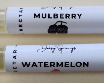 May include: Two clear nail oil pens. One has a label that reads "Mulberry" with a black berry illustration. The other pen has a label that reads "Watermelon". Both pens have white caps and are filled with a yellow liquid.