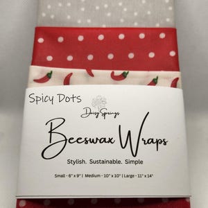 May include: A stack of beeswax wraps with a variety of patterns. The wraps are in shades of red, white, and gray, with polka dots and chili pepper designs. The packaging reads "Spicy Dots Beeswax Wraps" and lists sizes: Small - 6" x 9", Medium - 10" x 10", and Large - 11" x 14".