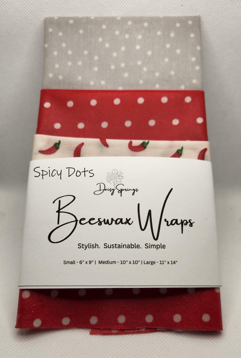 May include: A stack of beeswax wraps with a variety of patterns. The wraps are in shades of red, white, and gray, with polka dots and chili pepper designs. The packaging reads "Spicy Dots Beeswax Wraps" and lists sizes: Small - 6" x 9", Medium - 10" x 10", and Large - 11" x 14".