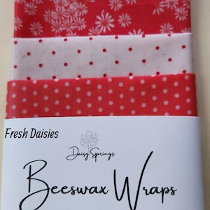 May include: A stack of red and white beeswax wraps with floral and polka dot patterns. The label reads "Fresh Daisies" and "Beeswax Wraps". Sizes are small (6" x 9"), medium (10" x 10"), and large (11" x 14").
