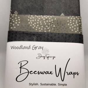 May include: A stack of gray beeswax wraps with various patterns, including trees and polka dots. The label reads "Woodland Gray Beeswax Wraps" by Daisy Springs. Sizes include small (6" x 9"), medium (10" x 10"), and large (11" x 14").