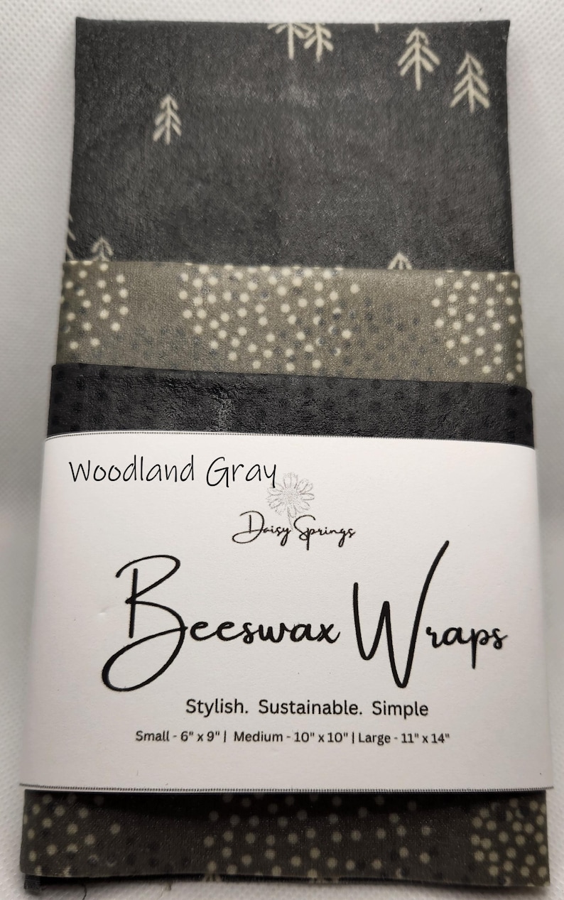 May include: A stack of gray beeswax wraps with various patterns, including trees and polka dots. The label reads "Woodland Gray Beeswax Wraps" by Daisy Springs. Sizes include small (6" x 9"), medium (10" x 10"), and large (11" x 14").
