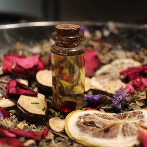 May include: A small clear glass vial filled with a golden liquid and sealed with a brown cork stopper. The vial is surrounded by a mixture of dried red rose petals, purple flowers, sliced dried citrus wheels, and small woody botanical pieces.