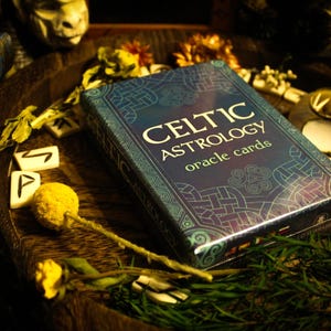 May include: A rectangular deck of Celtic Astrology oracle cards in a blue box with intricate knotwork patterns. The text on the box reads Celtic Astrology oracle cards. The deck is surrounded by small white tiles with black symbols and yellow flowers.