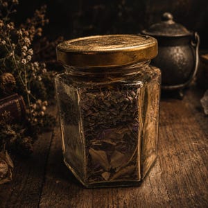 May include: A hexagonal glass jar with a metallic gold lid, filled with a mixture of dried herbs and botanical elements. The clear glass container features six flat sides and a wide mouth, showcasing the textured contents inside the vessel.