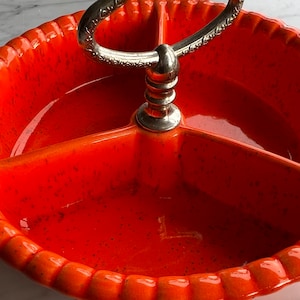 Op de afbeelding: Oranje keramische serveerschaal met drie secties en een zilveren metalen handvat. De schaal heeft een gespikkelde glazuur.