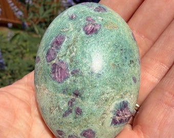 May include: A polished, oval-shaped gemstone with a mottled teal green base color. The stone is speckled with irregular patches of deep magenta-colored minerals. The smooth surface of the stone reflects light, highlighting the contrasting colors and textures.