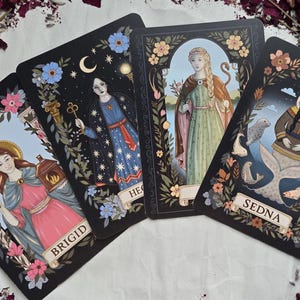 May include: Four tarot cards with illustrations of female figures. The cards feature floral borders and the names "BRIGID", "HECATE", and "SEDNA". The artwork includes celestial elements, keys, and a serpent, with a color palette of black, blue, pink, and gold.