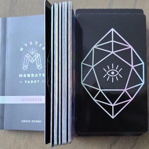 May include: A tarot deck set with a guidebook. The guidebook is gray with the text "Mystic Mondays Tarot" and "Guidebook". The deck of cards has a black background with a white geometric design and an eye symbol.