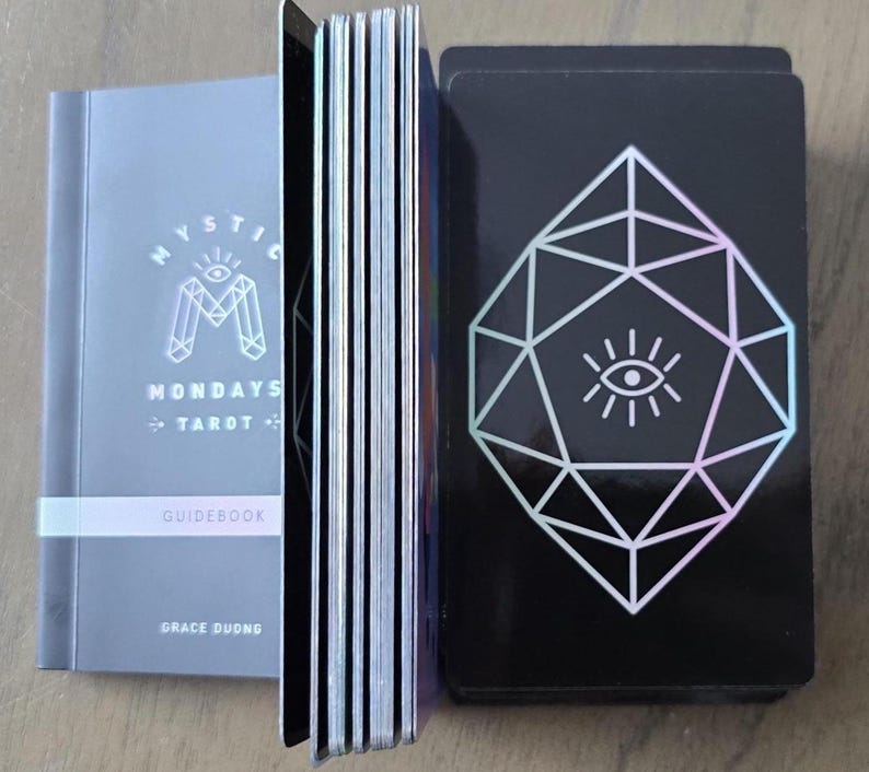 May include: A tarot deck set with a guidebook. The guidebook is gray with the text "Mystic Mondays Tarot" and "Guidebook". The deck of cards has a black background with a white geometric design and an eye symbol.