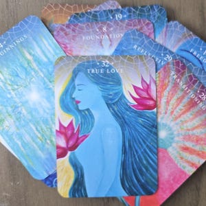 May include: A set of colorful oracle cards with various images and numbers. The cards feature a blue woman with long hair, a pink lotus flower, and a rainbow-colored background. The cards are numbered 14, 19, 24, 28, 29, 32, and feature words like "Freedom", "Foundation", "New Beginnings", "Transitioning Worlds", "Reflective", and "True Love".