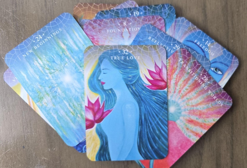 May include: A set of colorful oracle cards with various images and numbers. The cards feature a blue woman with long hair, a pink lotus flower, and a rainbow-colored background. The cards are numbered 14, 19, 24, 28, 29, 32, and feature words like "Freedom", "Foundation", "New Beginnings", "Transitioning Worlds", "Reflective", and "True Love".